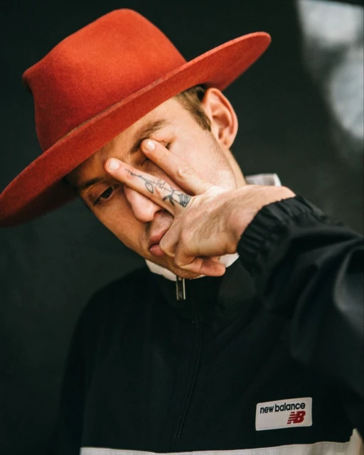 Close-up of a person wearing an orange hat and a black and white jacket, posing with fingers near the eye against a dark background.