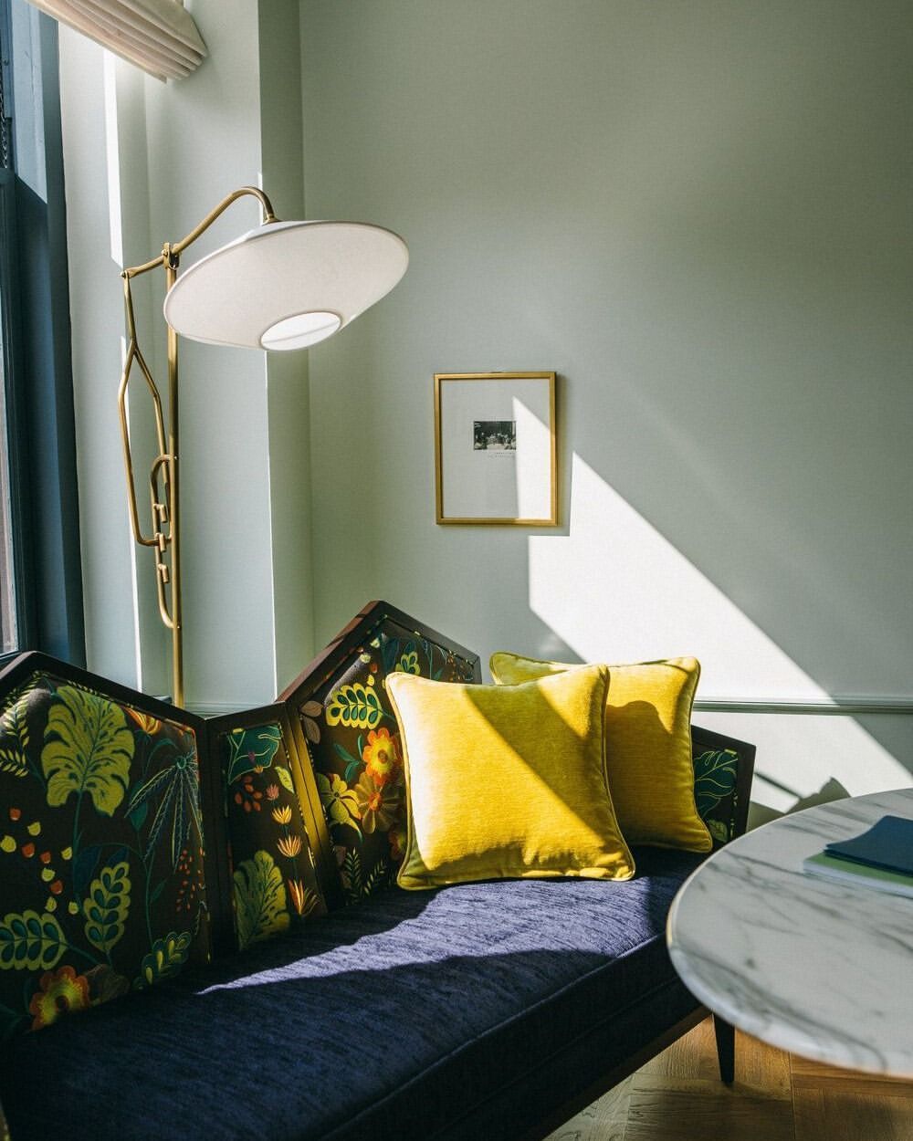 Seating area with blue couch, yellow pillows, floor lamp, and round marble table. The room is lit by natural window light.
