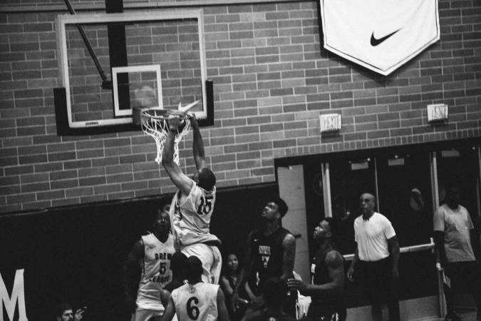 Action shot of a basketball player dunking during a game, surrounded by other players on a brick-walled indoor court with a Nike banner.
