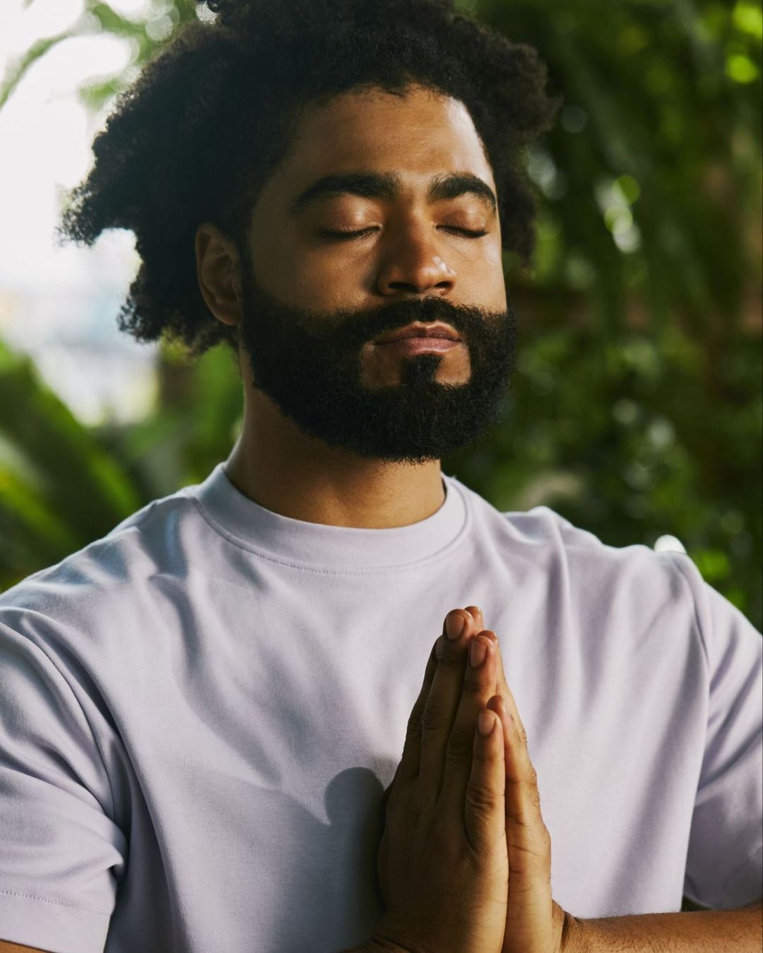 Adult male with afro and beard in light purple shirt with hands together in prayer or meditation outdoors among green foliage.