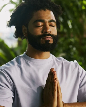 Adult male with afro and beard in light purple shirt with hands together in prayer or meditation outdoors among green foliage.