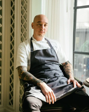 Portrait of a bald man with tattooed arms wearing a chef coat and dark apron, seated indoors.