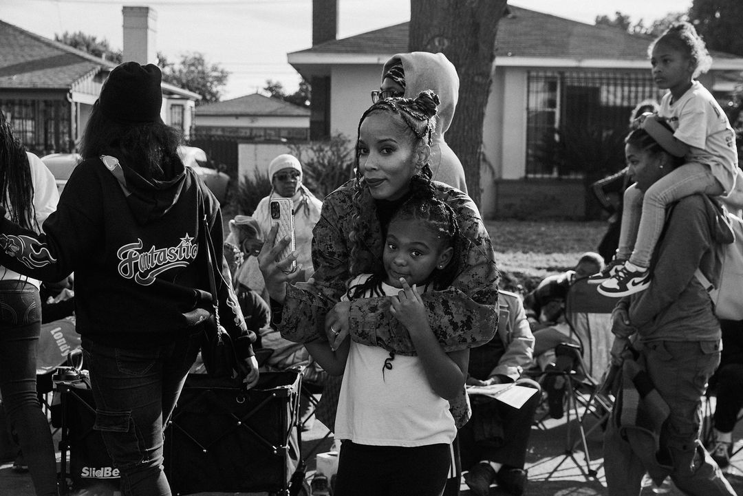 Black and white outdoor shot of a mother and daughter embraced, surrounded by a group of people in a residential street setting.