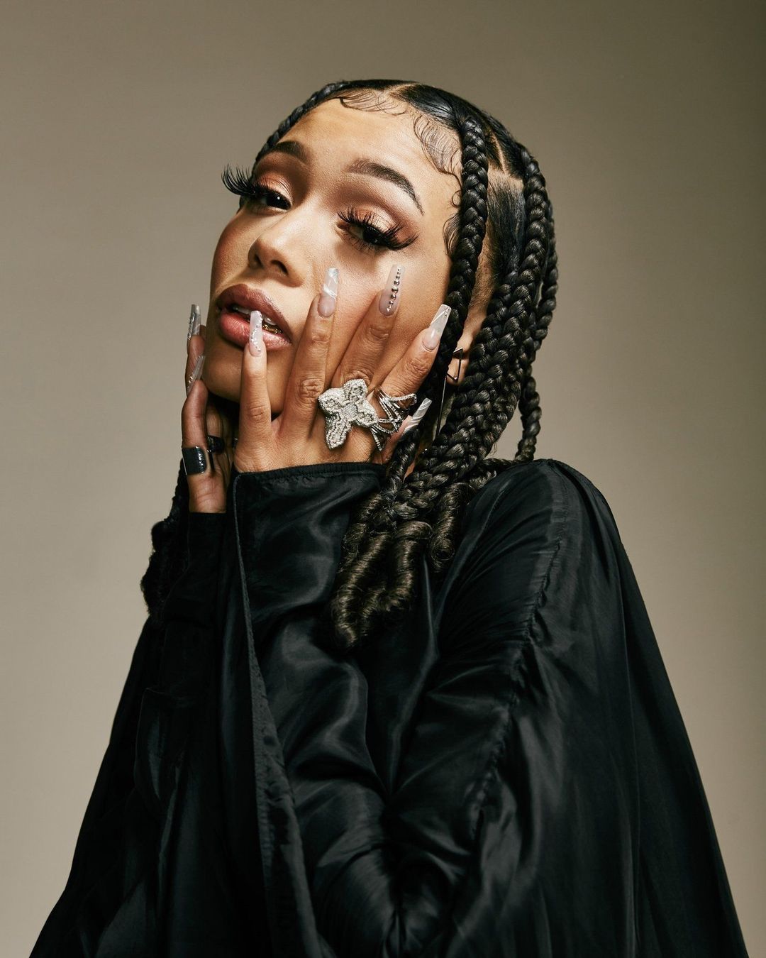 Close-up portrait of a young adult woman with braided hair, wearing a black jacket and rings, set against a light beige background.