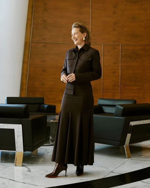 Elegant adult woman in a brown skirt and jacket smiles while standing indoors near black armchairs and a brown paneled wall.