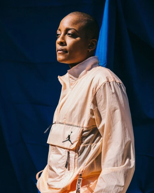 A fashionable woman with a shaved head, wearing a peach-colored jacket with black logo, stands against a blue backdrop in soft light.