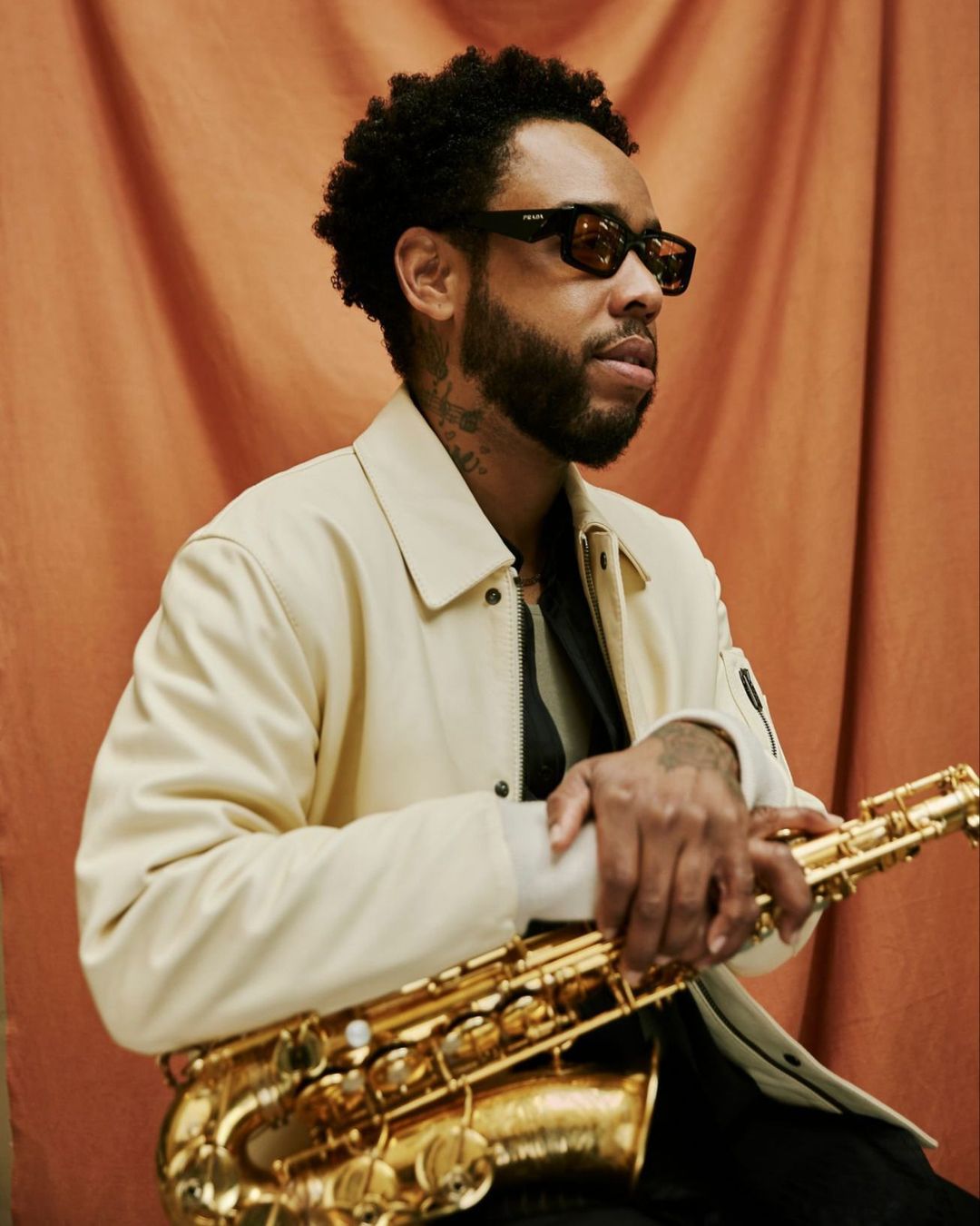 Adult male holding a saxophone in front of a rust-colored backdrop. The man is wearing sunglasses and a light-colored jacket.