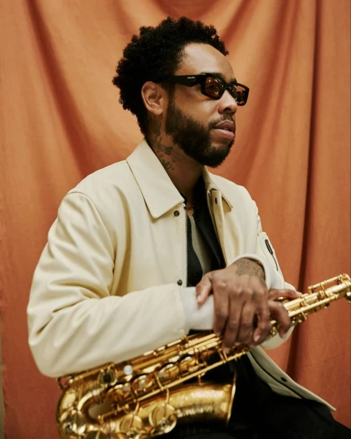 Adult male holding a saxophone in front of a rust-colored backdrop. The man is wearing sunglasses and a light-colored jacket.