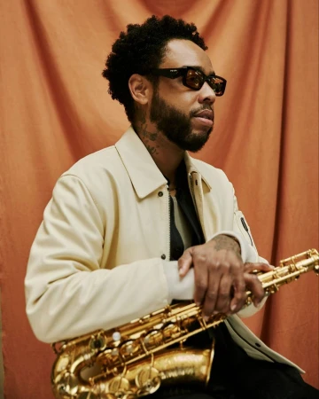 Adult male holding a saxophone in front of a rust-colored backdrop. The man is wearing sunglasses and a light-colored jacket.