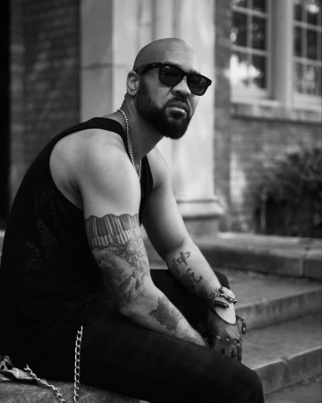 Black and white portrait of a bald man with a beard wearing sunglasses and a black tank top seated outdoors on stone steps