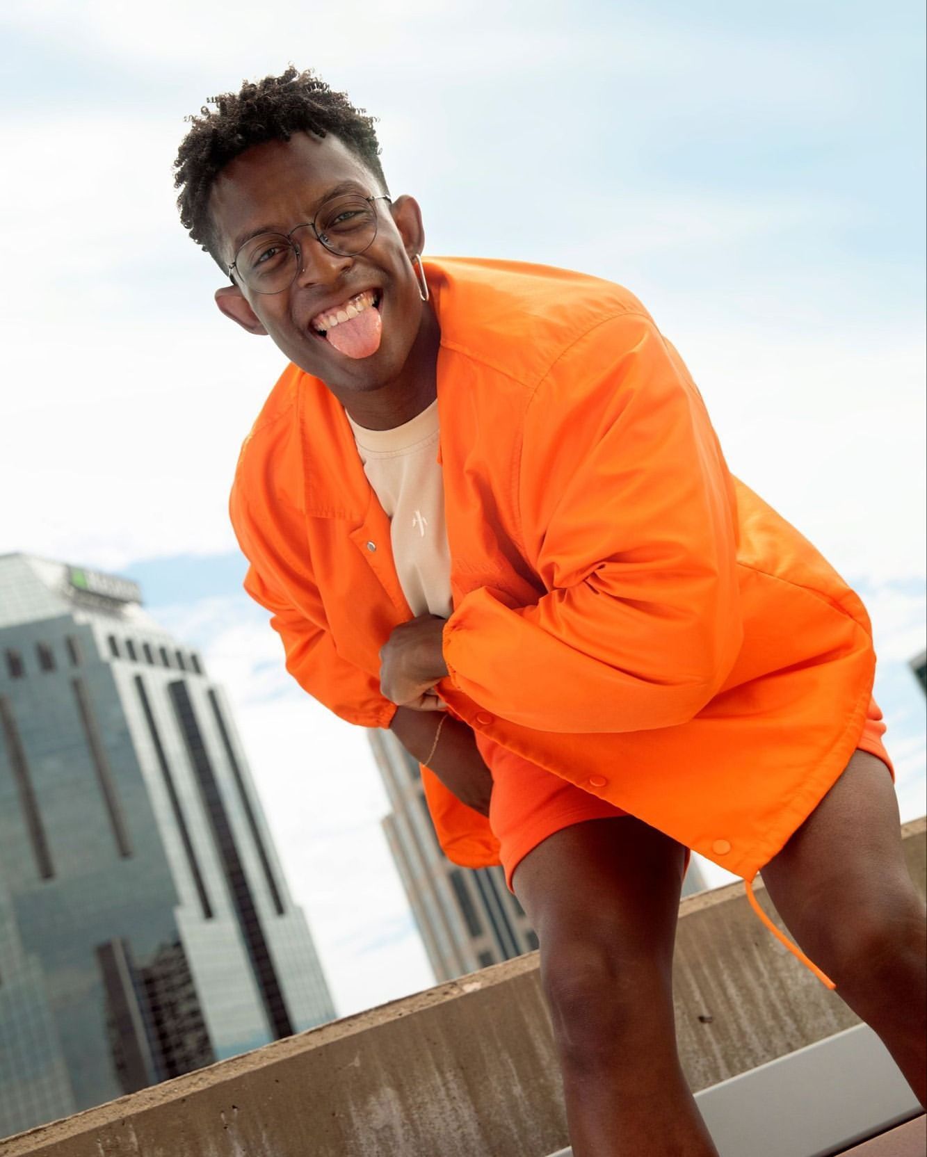 A young adult man poses outdoors with a playful expression, wearing an orange jacket and shorts with city buildings in the background.