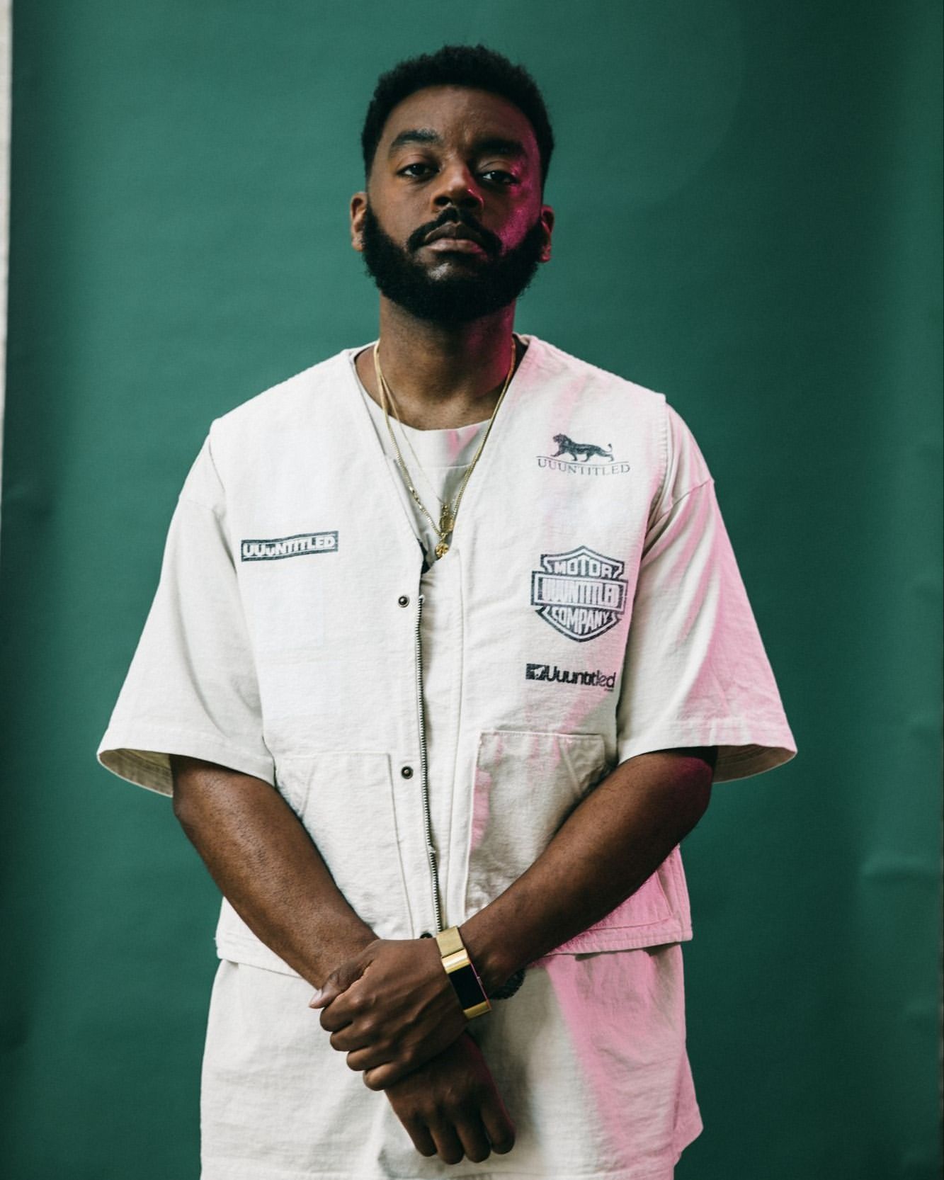 Portrait of an adult man with a beard in a cream-colored outfit in front of a green backdrop, wearing jewelry and a gold watch.