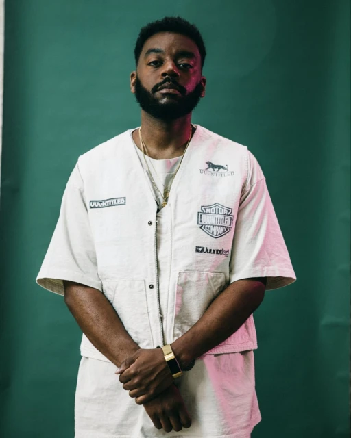 Portrait of an adult man with a beard in a cream-colored outfit in front of a green backdrop, wearing jewelry and a gold watch.