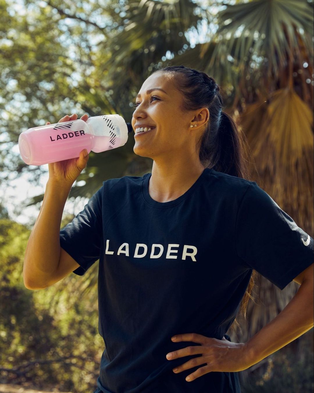 Adult female drinks from branded water bottle wearing a black t-shirt in a park setting with palm trees under natural daylight.