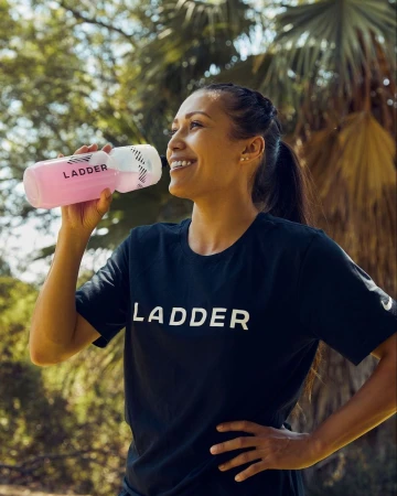 Adult female drinks from branded water bottle wearing a black t-shirt in a park setting with palm trees under natural daylight.