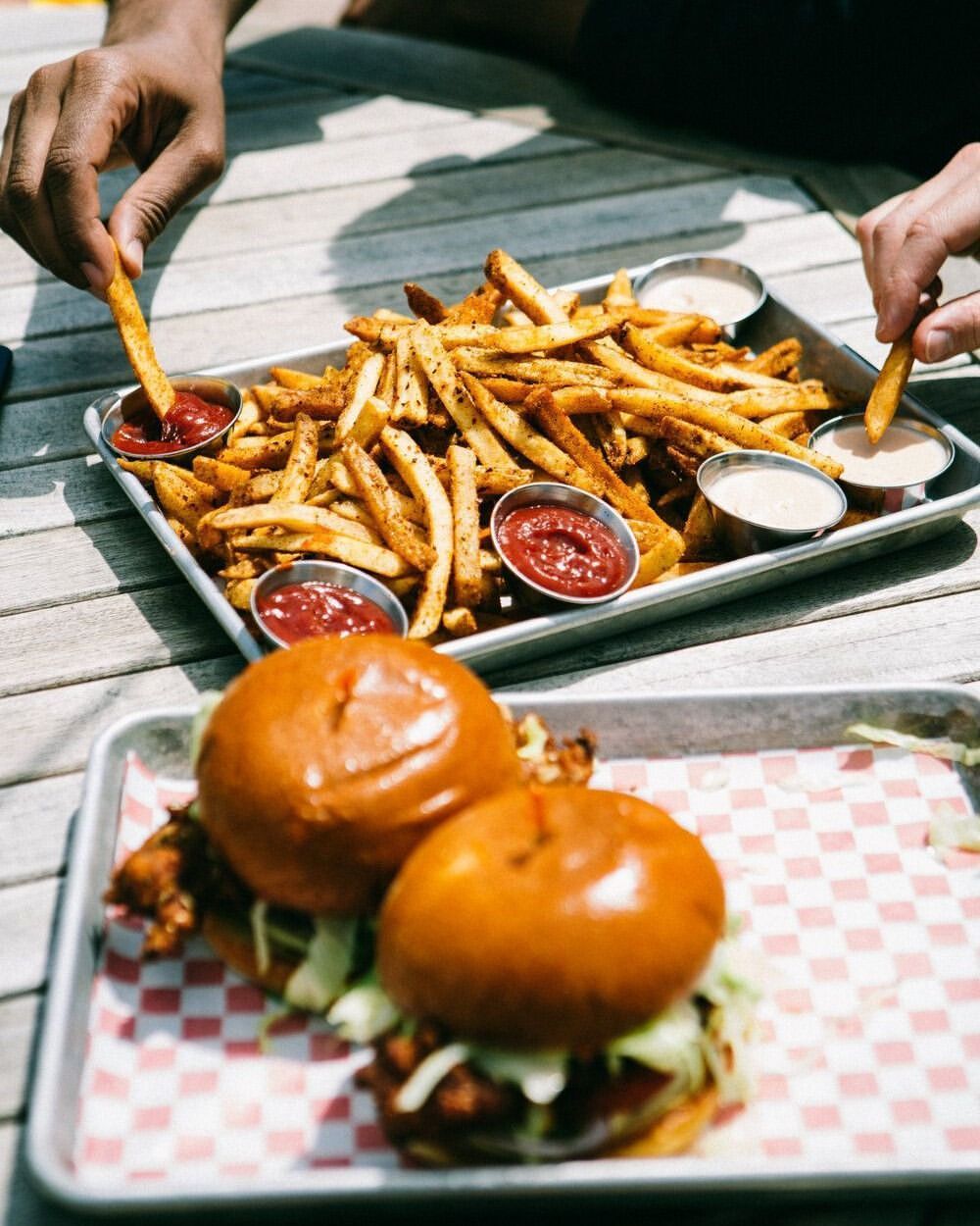 Overhead shot of two chicken sandwiches and a tray of french fries served on a wooden table, with two people eating.