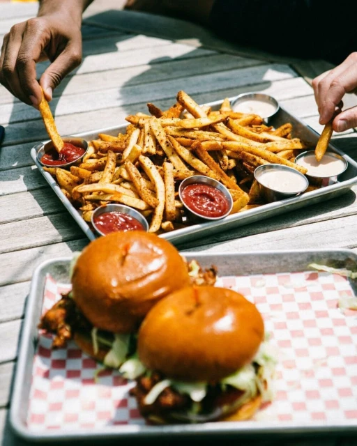 Overhead shot of two chicken sandwiches and a tray of french fries served on a wooden table, with two people eating.