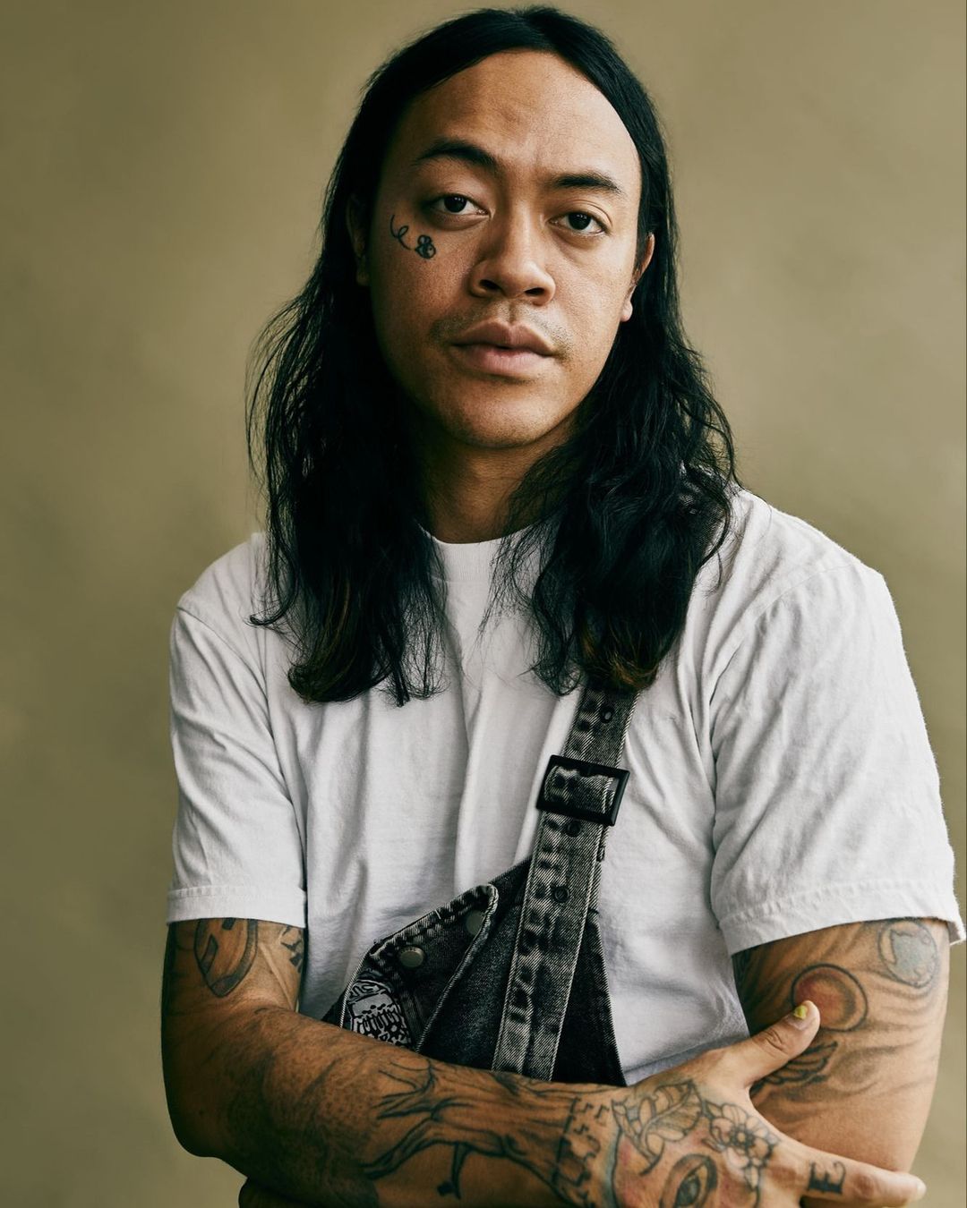 Portrait of a man with long hair and tattoos wearing a white t-shirt and dark denim overalls, captured in a close-up shot against a tan background.