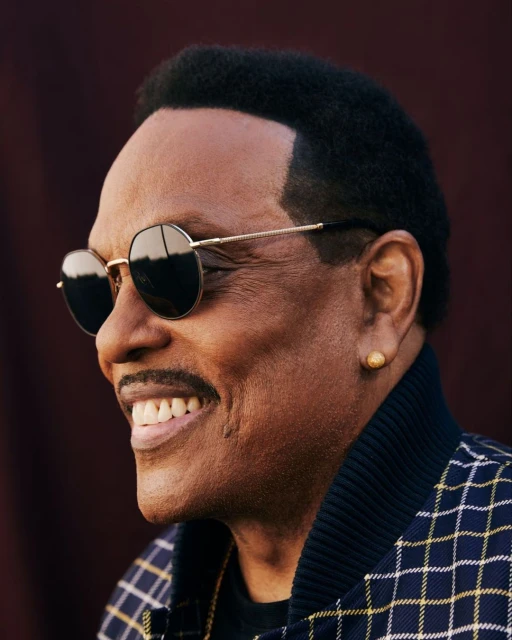 Smiling African American man in round sunglasses and a patterned jacket, posing in front of a blurred burgundy background in a close-up shot.