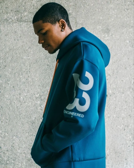 Portrait of a young adult male wearing a blue hooded sweatshirt, against a neutral concrete background. Features '23 ENGINEERED' text on the sleeve.