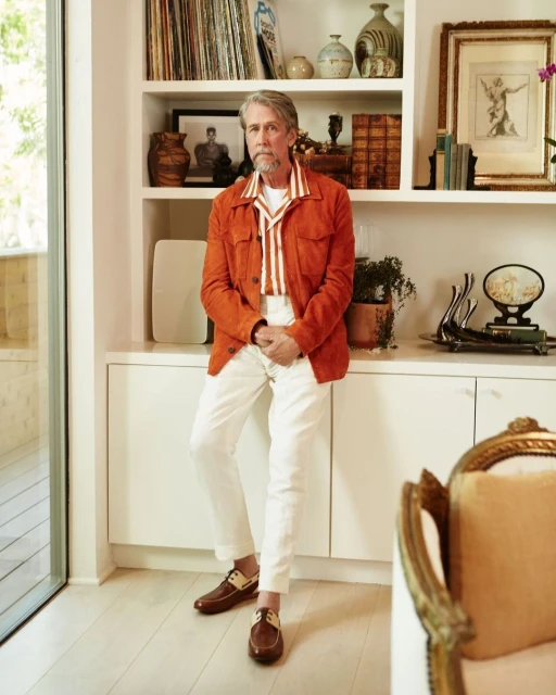 Indoor portrait of an adult male with grey hair, wearing an orange jacket, white pants, and brown shoes, posing near a white shelf and couch.