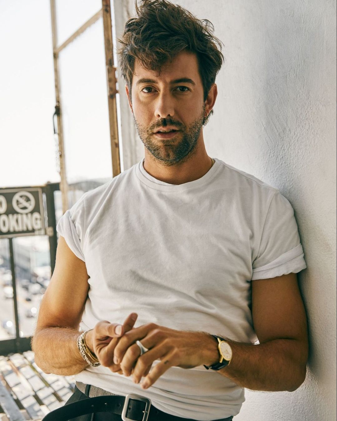 Adult male in a white t-shirt leans against a wall with a 'No Smoking' sign visible outdoors in an urban setting, portrait orientation.