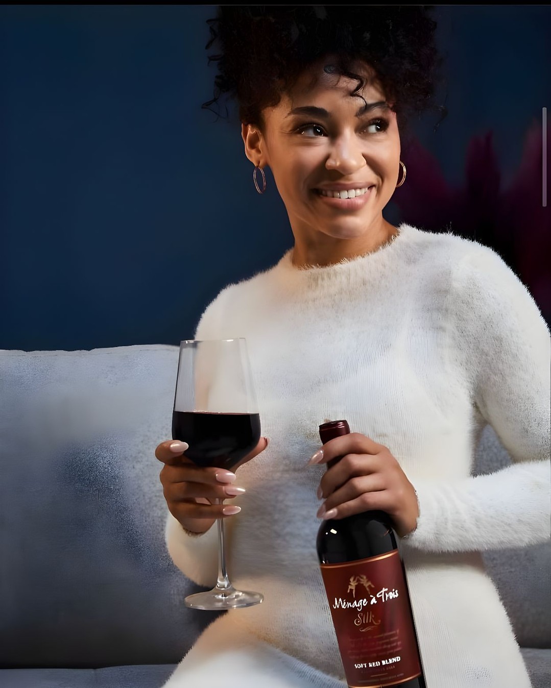 Smiling woman in white sweater poses with glass and bottle of Menage a Trois Silk Red Blend, seated against a dark blue background.