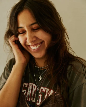Close-up portrait of a smiling woman wearing layered necklaces and a graphic t-shirt against a plain wall background