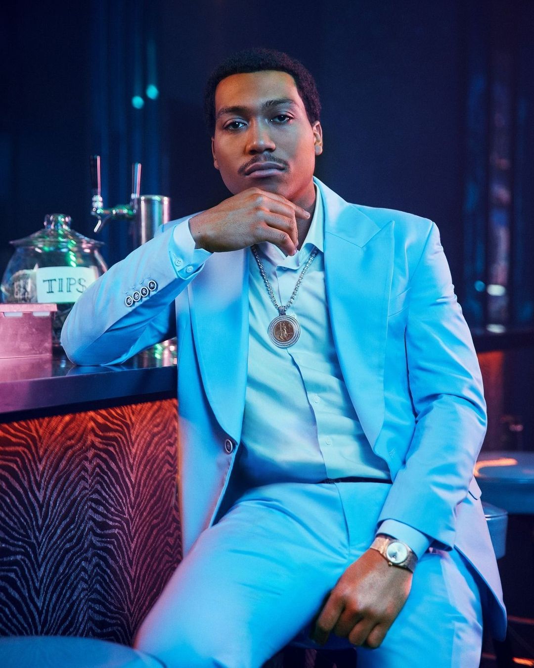 Man in an aqua suit poses in front of a bar, showcasing a vintage aesthetic with a gold watch and necklace, complemented by mood lighting.