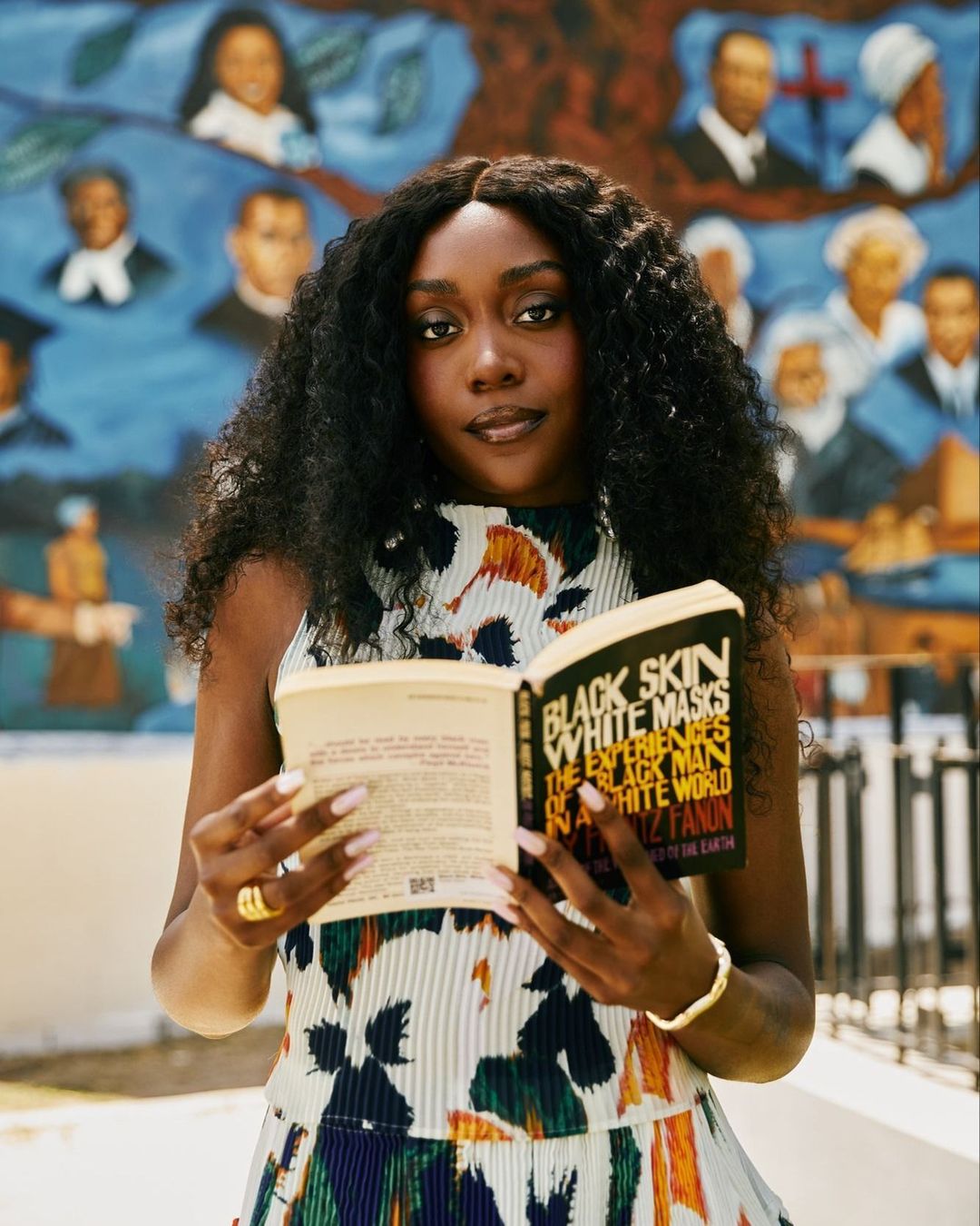 A stylish adult female with dark curly hair reads 'Black Skin White Masks' by Frantz Fanon, posing in front of a colorful mural.