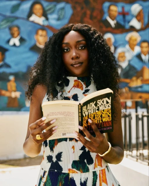 A stylish adult female with dark curly hair reads 'Black Skin White Masks' by Frantz Fanon, posing in front of a colorful mural.
