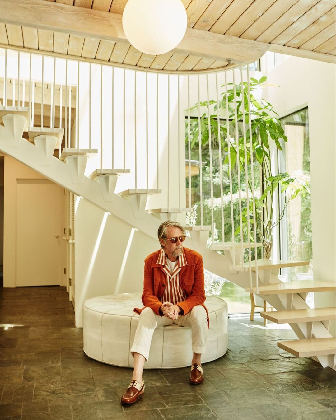 Man in an orange jacket and sunglasses sits on a round ottoman in a modern home interior with a winding staircase and plant.