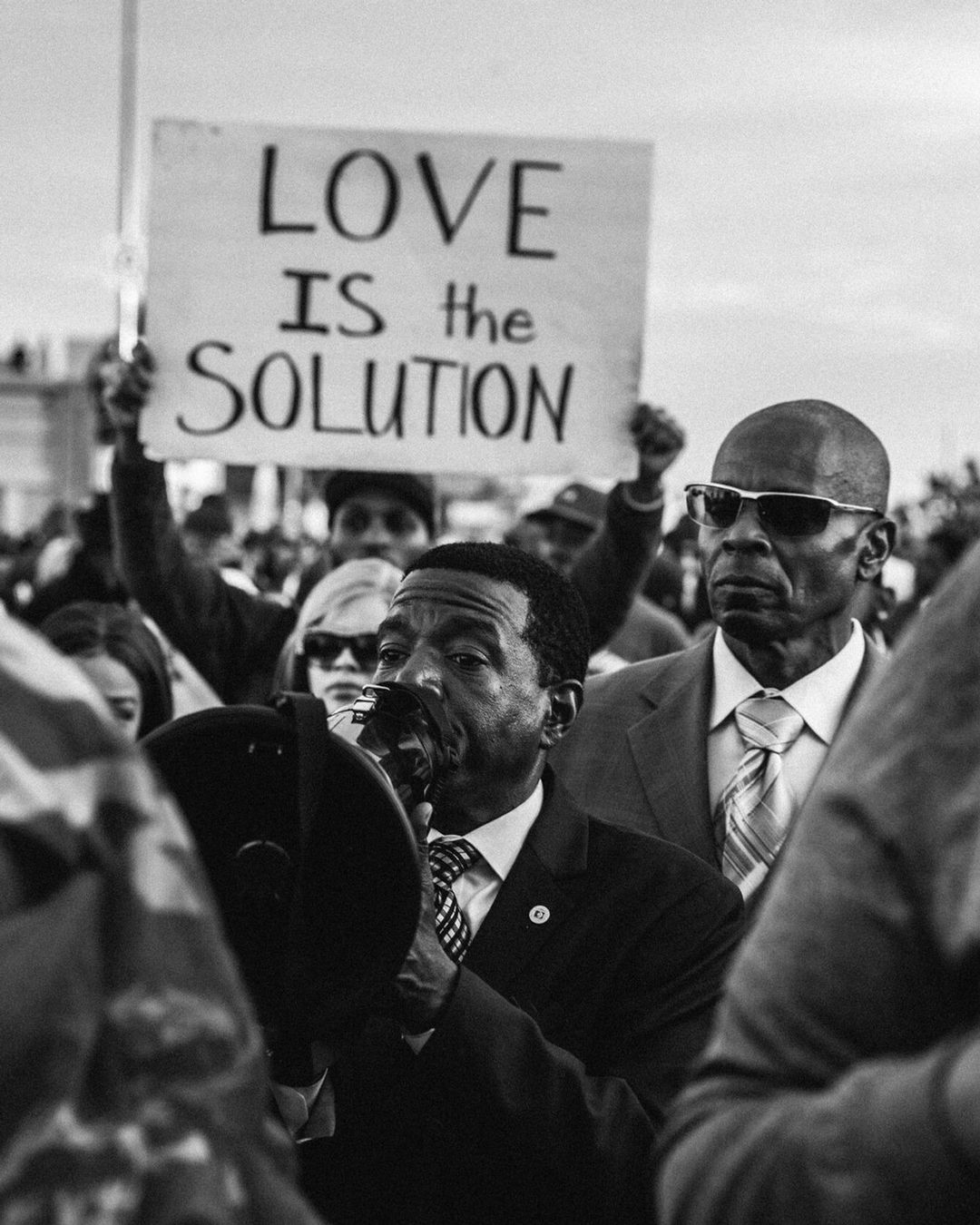 Black and white photograph of a group of people at a rally, an adult male with a bullhorn, holding sign stating 'Love is the Solution'.