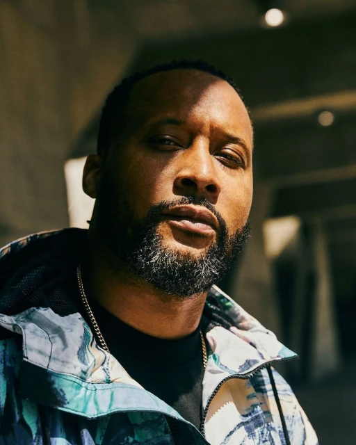 Portrait of a man with a beard wearing a patterned jacket stands against a concrete structure in natural light.