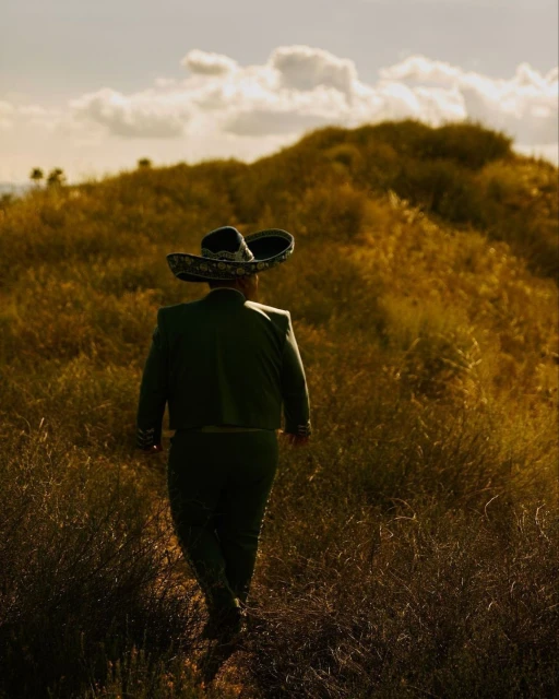 Man in charro suit walks across a golden field beneath a partly cloudy sky. Wide-brimmed sombrero shades him from behind.