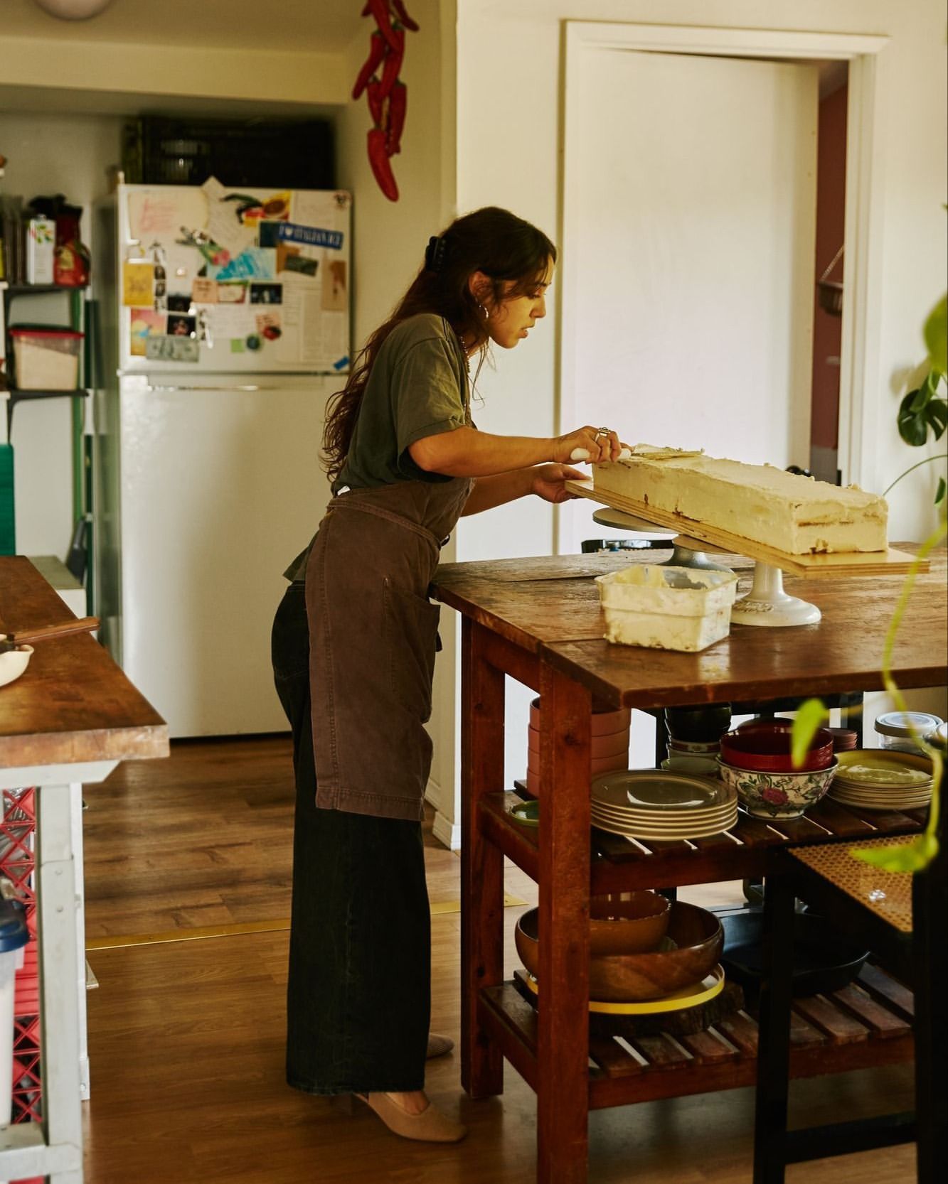 A woman in an apron standing in a kitchen, frosting a rectangular cake with white icing on a wooden table with shelves.