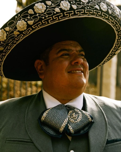 Man wearing a grey suit and a black sombrero smiles outdoors. The traditional Mexican hat features floral embroidery along the brim.
