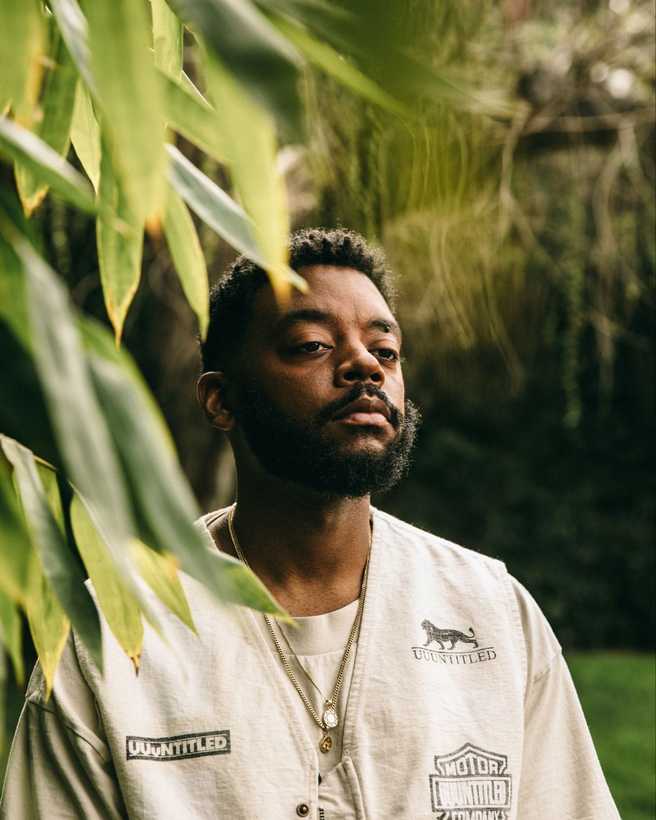 Portrait of a bearded Black male wearing layered gold necklaces and vest with foliage framing the shot in an outdoor setting.