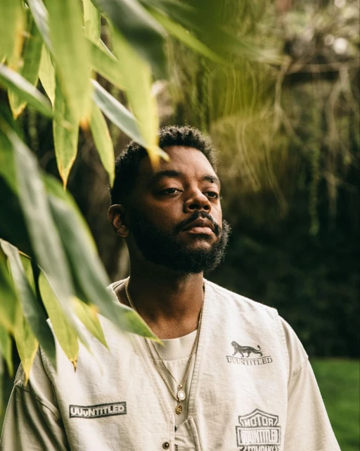 Portrait of a bearded Black male wearing layered gold necklaces and vest with foliage framing the shot in an outdoor setting.