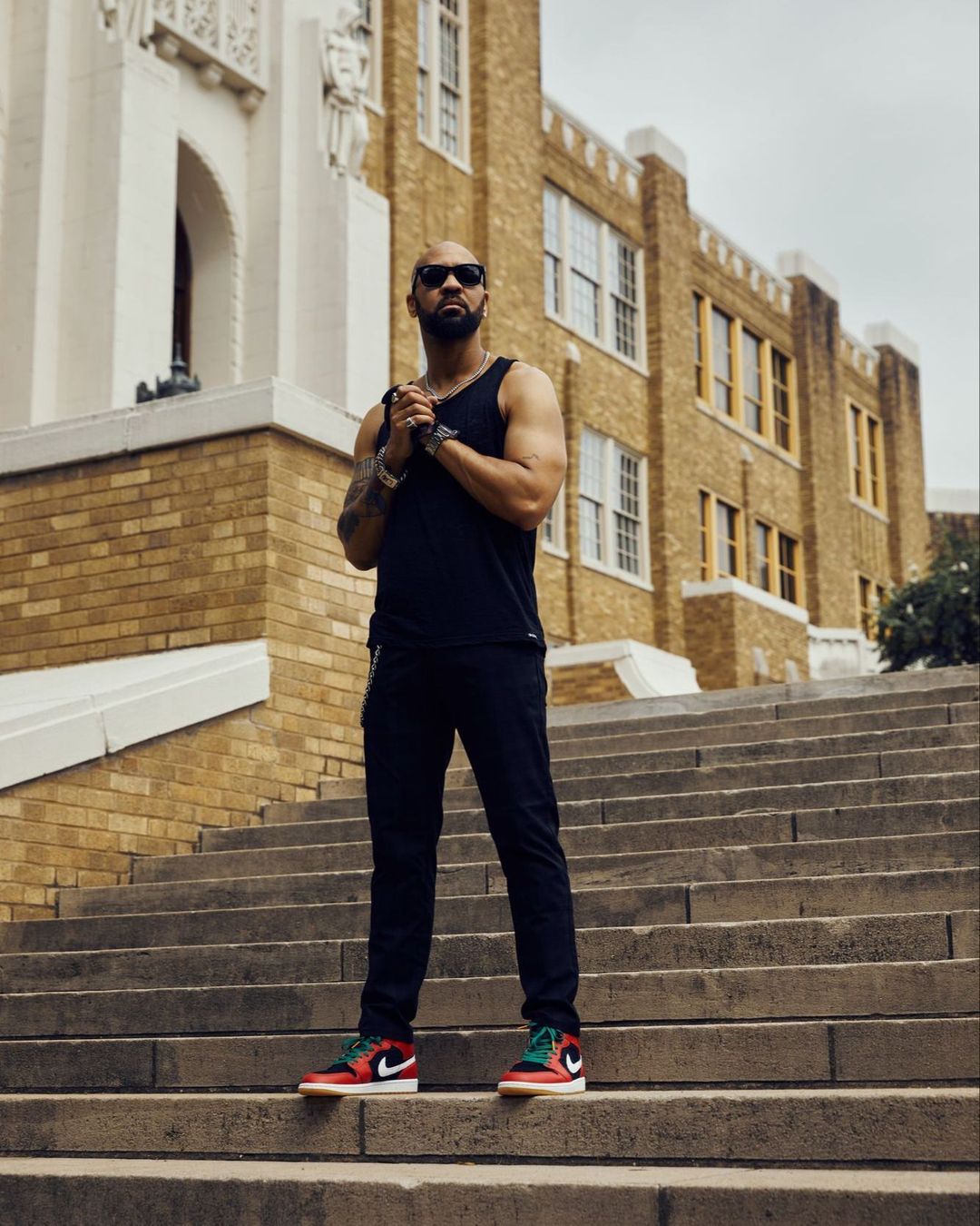 Stylish bald man with sunglasses and black tank top poses on concrete steps of brick building with classic red and green sneakers.