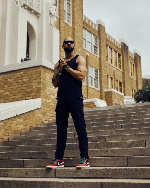 Stylish bald man with sunglasses and black tank top poses on concrete steps of brick building with classic red and green sneakers.