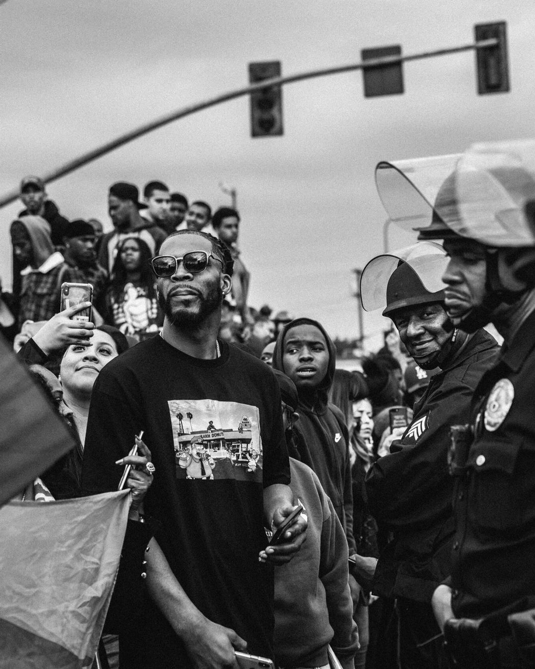 A crowd of people including a man in sunglasses and a t-shirt with an image, standing in front of police officers with shields in a black and white photograph.