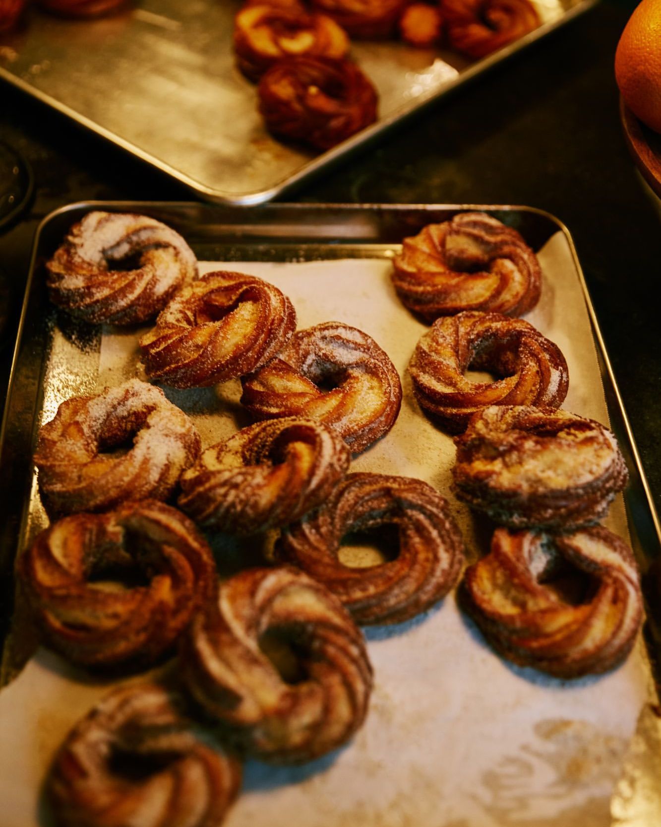 Several trays of freshly baked kouign-amann pastries with a golden-brown color, some dusted with sugar, arranged indoors.