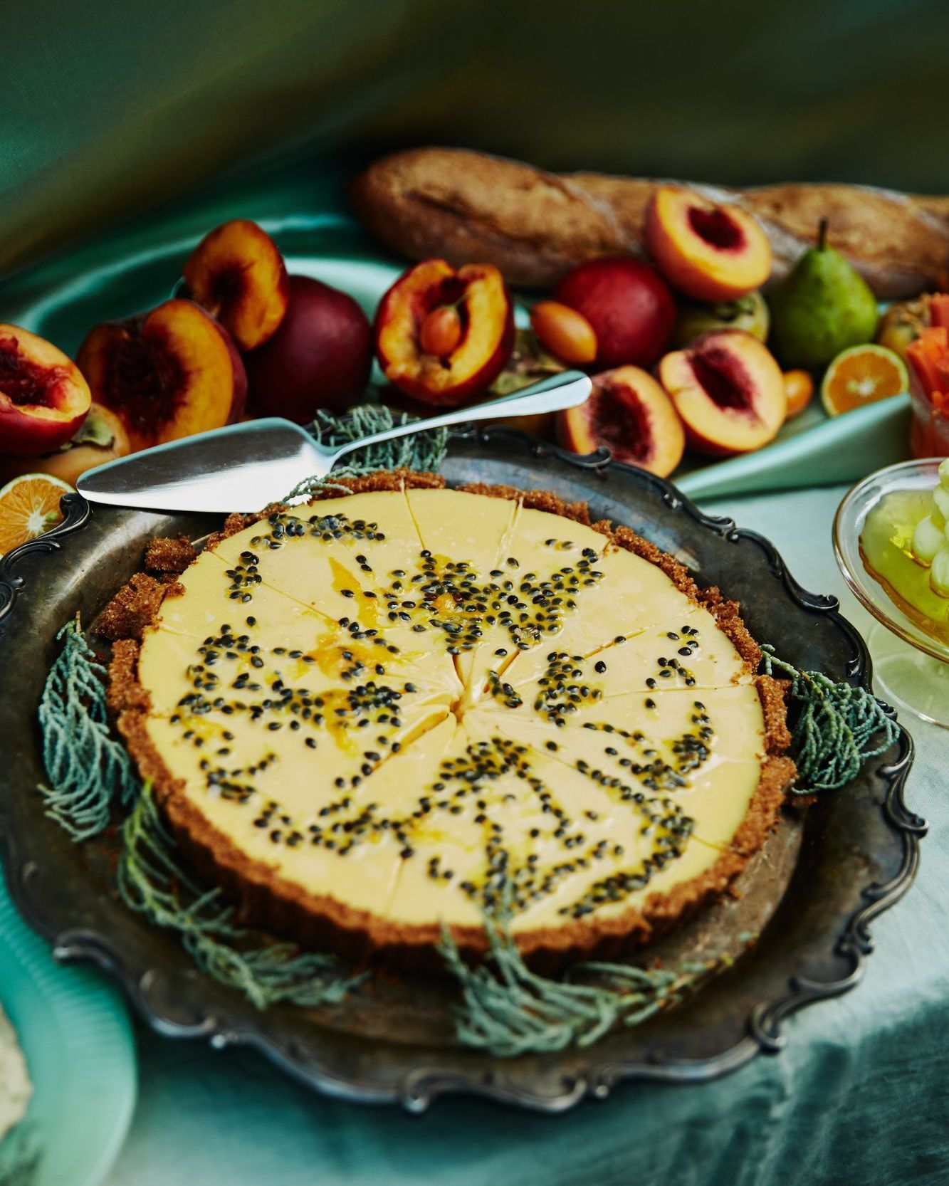 Overhead shot of a passionfruit tart on a silver platter, surrounded by assorted fresh fruits and a baguette. Still life, indoor lighting.