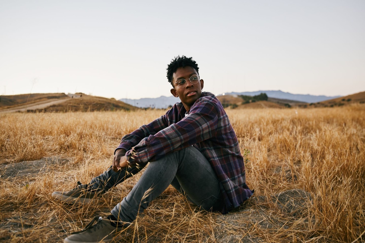 Man with glasses in a plaid shirt sitting in a field of dry grass in the sunlight, with hills and the sky in the background.