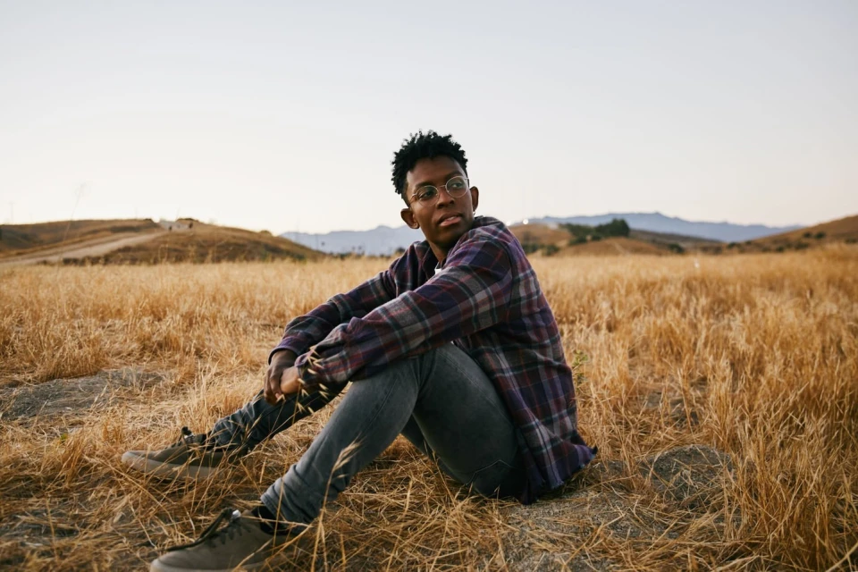 Man with glasses in a plaid shirt sitting in a field of dry grass in the sunlight, with hills and the sky in the background.