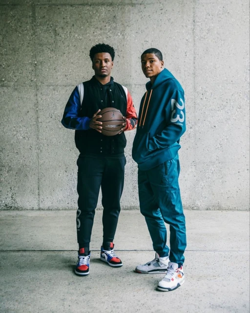 Two young men pose in athletic wear, one holding a basketball, against a textured concrete wall in a medium shot.