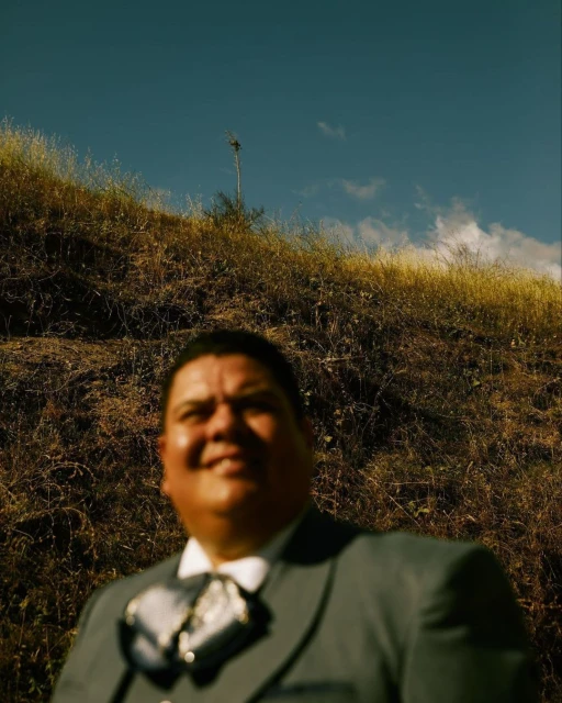 An adult man in formal wear smiles at the camera in front of a hillside with a clear blue sky in the background.