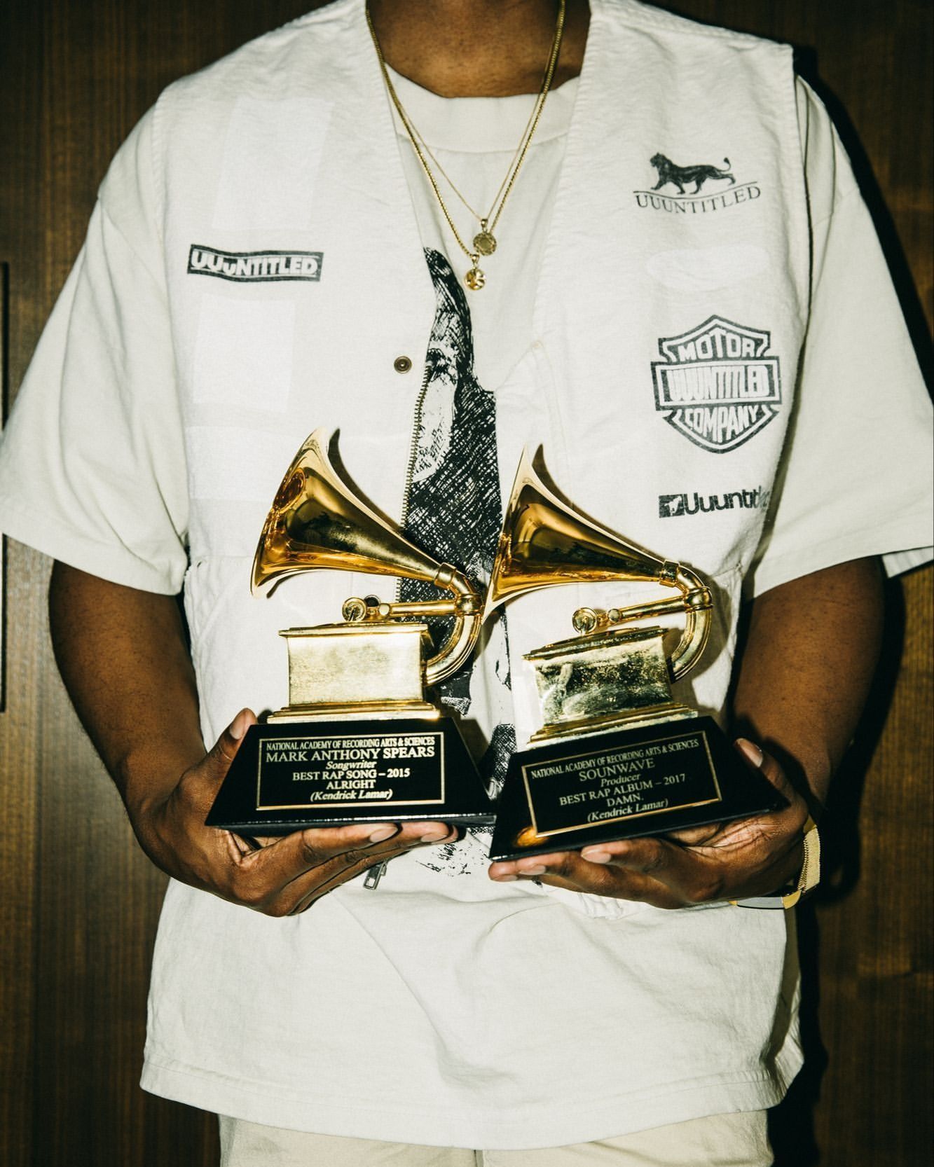 Man holding two gold Grammy awards, wearing a white t-shirt and vest, standing indoors with a wood panel background.