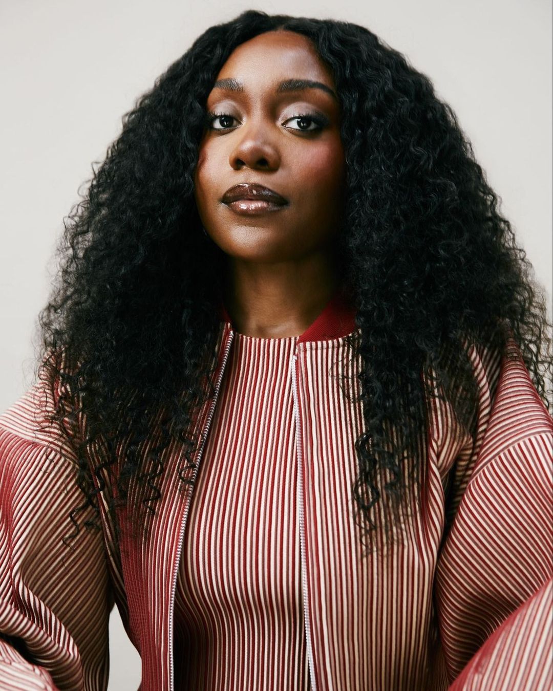 Portrait of a dark-skinned woman with curly black hair, wearing a striped jacket, set against a neutral background.
